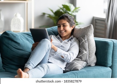 Relaxed indian girl lying on comfortable couch in cozy light living room holding tablet wireless gadget smiling enjoy remote chat with family by video call application. Web shop happy customer concept - Powered by Shutterstock