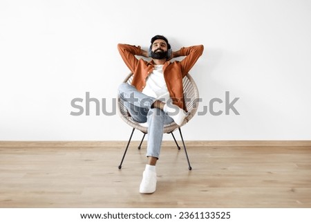 Relaxed Indian Young Man With Wireless Headphones Resting And Listening To Music On Weekend, Sitting In Chair With Hands Behind Head, Enjoying Break And Relaxation Time At Home