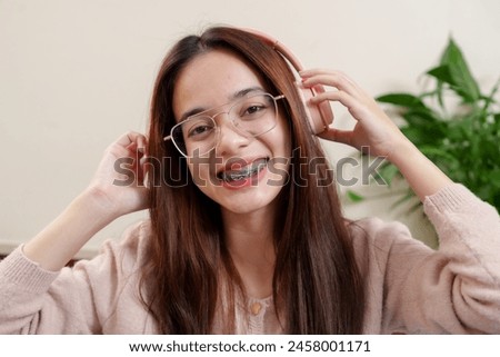 Relaxed individual with headphones, adjusting with one hand, cheerful smile, teeth with braces shown, casual attire, indoor comfort, foliage in blurred backdrop. Holiday happiness Asian teenagers