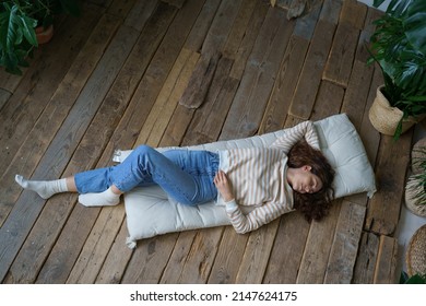 Relaxed Italian woman lying on wooden floor at home, rest after work. Top view of calm young Spanish female sleeping on mattress surrounded by tropical houseplants in greenhouse. Wellness concept. 