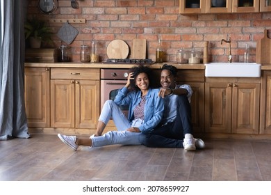 Relaxed Joyful Young African American Family Couple Sitting On Warm Heated Wooden Floor In Stylish Cottage House Kitchen, Spending Weekend Time In Own Dwelling, Real Estate Concept.