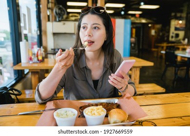 Relaxed Korean woman enjoying brunch and using phone at same time is looking at scenery outside the window of eatery. Hungry female visitor taking a bite of food with phone in hand in carefree mood.