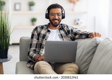 Relaxed Long-haired, Bearded Middle-eastern Guy Watching Movie Online, Reclining On Couch And Using Laptop And Headset In Living Room. Positive Arab Man Having Video Call Via Notebook, Copy Space