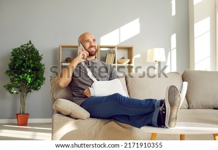 Relaxed man with broken arm having fun talking on phone with friends during rehabilitation at home. Cheerful bald young man in immobilizer on injured arm sitting on sofa throwing legs on coffee table.