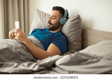 Relaxed man chilling in bed with cellphone and wearing wireless headphones, watching movie, videos or TV show on Internet, sitting in bed, copy space - Powered by Shutterstock