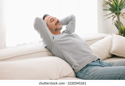 Relaxed man enjoying rest lying on comfortable couch with the hands on the head in living room at home. Calm middle-age man breathing fresh air enjoying no stress free peaceful weekend in house. - Powered by Shutterstock