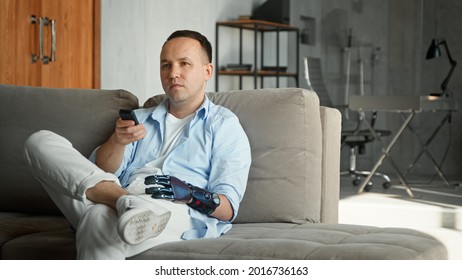 Relaxed Man With High Tech Bio Hand Prothesis Takes Remote Controller And Switches Television Channels Sitting On Large Sofa At Home