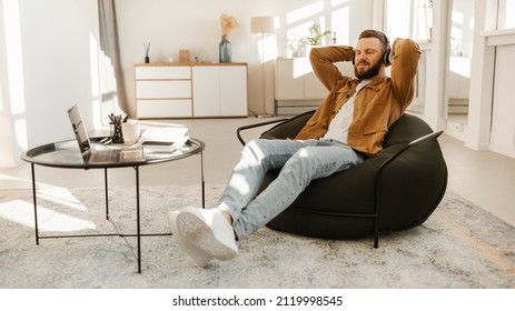 Relaxed Man Listening To Music Wearing Headphones Resting With Eyes Closed Sitting In Chair Holding Hands Behind Head At Home. Male Relaxes And Listens To Business Podcast Or Audiobook. Panorama