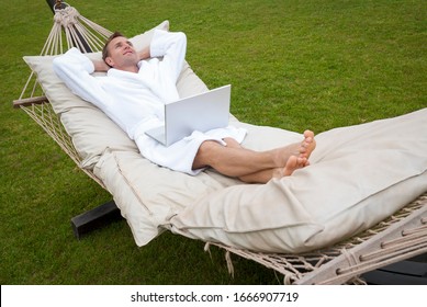 Relaxed Man In Plush White Robe Lying Back With Hands Behind His Head On A Large Outdoor Hammock With His Laptop Computer