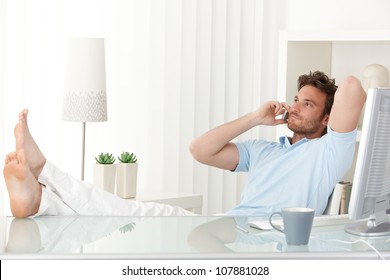 Relaxed man sitting with feet up on desk at home, talking on mobile phone, smiling.