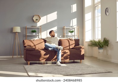 Relaxed man sitting on a comfortable couch at home. Happy young man enjoying free time and lounging on a soft brown sofa in a cosy living room interior with light grey walls, shelves and green plants - Powered by Shutterstock