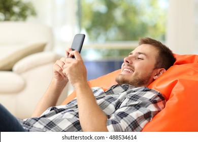 Relaxed man using a smart phone lying on an orange pouf in the living room at home with a window in the background - Powered by Shutterstock