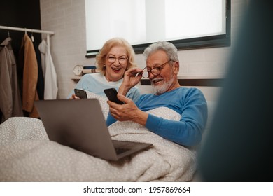 Relaxed Married Man And Woman Sitting In Bed. Old Husband And Senior Wife Holding The Mobile Phone. They Are Smiling And Looking At Devices.