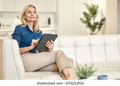 Relaxed Mature Lady In Casual Wear Looking Away, Using Tablet Pc While Sitting On A Couch In Modern Apartment On A Daytime