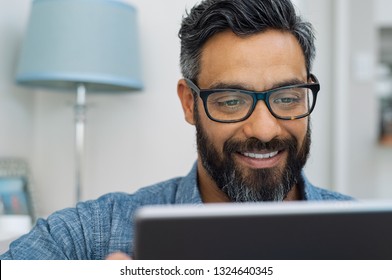 Relaxed Mature Man At Home Using Digital Tablet. Handsome Hispanic Man Using Laptop On Sofa. Confident Multiethnic Guy With Spectacles And Beard Using Digital Laptop.