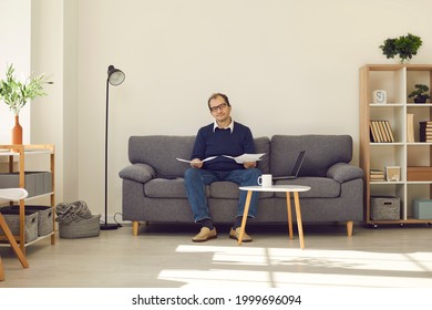 Relaxed mature man sitting on sofa in the living-room, holding papers and looking at camera. Senior person reading pension plan or going over business documents while doing paperwork at home