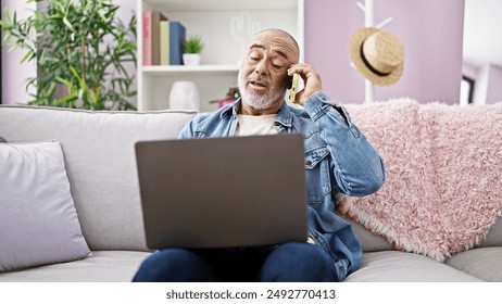 A relaxed mature man talks on the phone while working on a laptop in a cozy living room. - Powered by Shutterstock