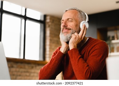 Relaxed Mature Middle-aged Man Businessman Freelancer Listening To The Music, Podcast, E-book, Radio Programme, Playlist, Musical Track In Headphones While Working Remotely On Laptop At Home