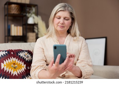 Relaxed Mature Old 60s Woman Middle Aged Female Holding Smartphone Using Mobile App, Texting Message, Search Ecommerce Offers On Cell Phone Technology Device Sitting On Couch At Home.