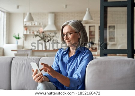 Relaxed mature old 60s woman, older middle aged female customer holding smartphone using mobile app, texting message, search ecommerce offers on cell phone technology device sitting on couch at home.