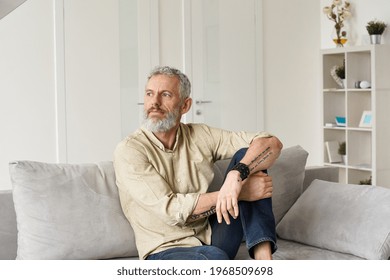 Relaxed Mature Older Thoughtful Tattooed Hipster Man Relaxing Sitting On Sofa And Thinking At Home. Pensive Calm Stylish Mid Aged Single Bearded Guy Resting On Couch At Home Looking Away, Reflecting.