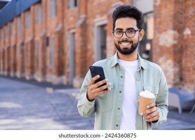 Relaxed middle eastern man holding telephone and plastic coffee cup while walking during daytime. Positive brunet freelancer with flexible work schedule taking break for beverage and scrolling feed. - Powered by Shutterstock