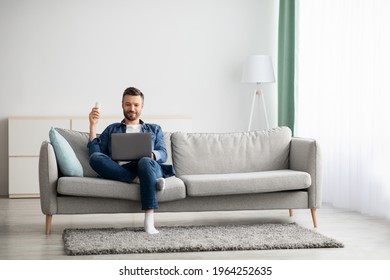 Relaxed middle-aged man with laptop sitting on couch at home, surfing on Internet or watching movie, raising hand with AC remote up, copy space. Cooling systems for home, air conditioner concept