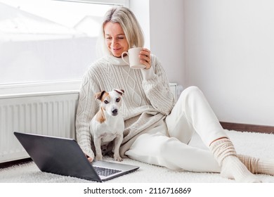 Relaxed Middle-aged Woman Is Working On A Laptop At Her Home. Adult Female Is Sitting On The Floor Near The Radiator In A White Knitted Sweater With Her Dog And Watching Movie
