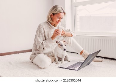 Relaxed Middle-aged Woman Is Working On A Laptop At Her Home. Adult Female Is Sitting On The Floor Near The Radiator In A White Knitted Sweater With Her Dog And Watching Movie
