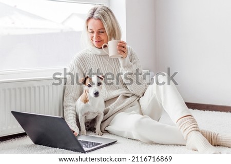 Relaxed middle-aged woman is working on a laptop at her home. Adult female is sitting on the floor near the radiator in a white knitted sweater with her dog and watching movie