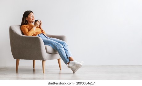 Relaxed Millennial Asian Woman Drinking Coffee, Having Break, Chilling In Armchair Against White Studio Wall, Panorama With Free Space. Lovely Young Lady Chilling And Enjoying Hot Drink