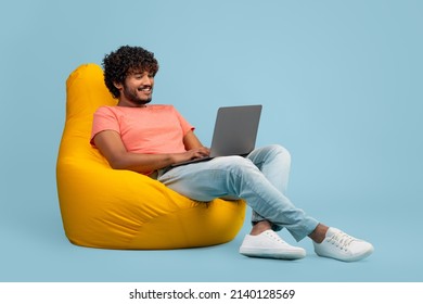 Relaxed millennial indian man in casual outfit sitting on bean bag, using modern laptop on blue studio background, typing on computer keyboard, playing video games, panorama with copy space - Shutterstock ID 2140128569