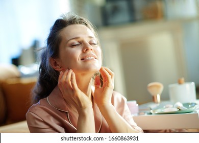 Relaxed Modern 40 Years Old Woman In Pajamas At Modern Home In Sunny Winter Day Applying Facial Cream And Making Massage.