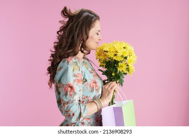 relaxed modern middle aged woman with long wavy brunette hair with yellow chrysanthemums flowers and green shopping bag against pink background. Foto Stock