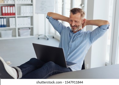 Relaxed modern successful businessman in jeans with his feet up on the desk in the office reading on a laptop computer with hands behind his head and smile of satisfaction