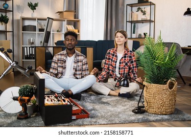 Relaxed Multi Ethnic Couple Sitting In Lotus Position And Meditating On Floor With Cardboard Boxes And Furniture Around. Young Man And Woman Taking Rest During Moving Process To New Apartment.