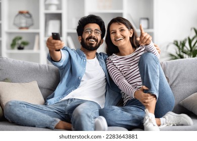 Relaxed multiracial spouses sitting on sofa at home, watching movie together, handsome middle eastern husband hugging his indian wife, enjoying TV show, holding remote control, copy space