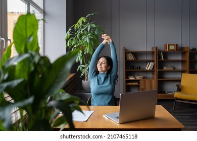 Relaxed office worker woman stretching hands and body taking break from work on laptop smiling look in window. Joyful freelancer copywriter girl happy with task done at workplace in coworking space