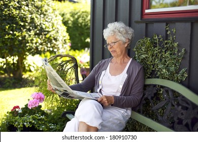 Relaxed Old Woman Sitting On Bench In Backyard Reading Newspaper