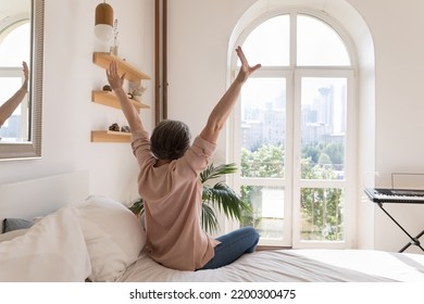 Relaxed Older Mature Woman Enjoying Being In Cozy Home Bedroom, Good Morning, Sitting On Bed, Feeling Great After Sleeping Enough On Comfortable Mattress, Rising Hands, Stretching Body. Back View