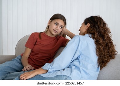 Relaxed optimistic woman and school age girl talking and sharing secrets sits on sofa indoors. Casual mom and teenage daughter with slight smile talk about important topics and discuss child friends - Powered by Shutterstock