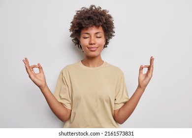 Relaxed peaceful curly haired young woman makes zen gesture keeps hads in mudra closes eyes practices yoga wears casual t shirt isolated over white background calms down during stressful day.