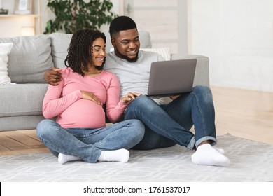 Relaxed pregnant african couple using laptop at home, sitting on floor at living room, enjoying weekend, copy space