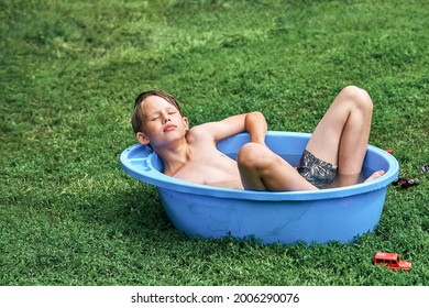 Relaxed Preteen Boy In Shorts Lies In Large Blue Plastic Basin With Water On Green Lawn Grass In Shady Yard Of Summerhouse