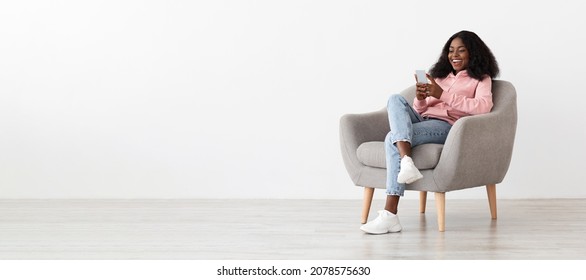 Relaxed pretty young black woman sitting in arm chair with cellphone at home, checking social media, empty white wall background, panorama with copy space. Gadgets addiction for millennials concept