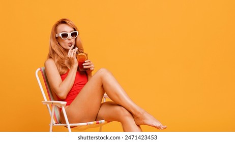 A relaxed red-haired woman in a red swimsuit and white sunglasses enjoys a cocktail while sitting on a striped beach chair against a vibrant orange background, embodying a carefree summer vibe. - Powered by Shutterstock