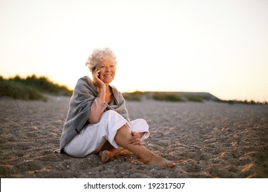 Relaxed retired woman wearing shawl sitting on sandy beach making a phone call. Old caucasian woman sitting on the beach looking at camera outdoors - Powered by Shutterstock