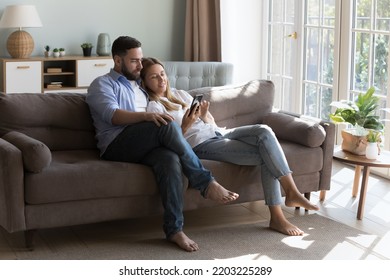 Relaxed Satisfied Millennial Couple Sharing Mobile Phone, Holding Cellphone, Resting On Couch, Hugging, Using Online App, Shopping On Internet Together, Talking. Family Communication Concept