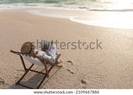Relaxed senior biracial woman reading book while sitting on folding chair at beach during sunset. lifestyle and hobbies.