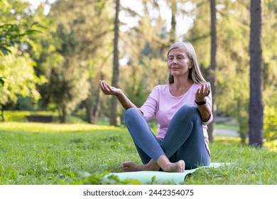 Relaxed senior gray-haired woman sitting in the park on a mat in the lotus position with closed eyes and meditating while doing yoga. - Powered by Shutterstock